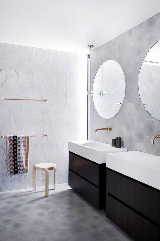 Bright contemporary bathroom with a double vanity, white rectangular basins, dark timber cabinetry, large oval mirrors, brass wall-mounted faucets and towel rails, pale marble-look wall panels, and grey hexagonal floor and feature wall tiles. A patterned towel and small stool add subtle decorative detail.
