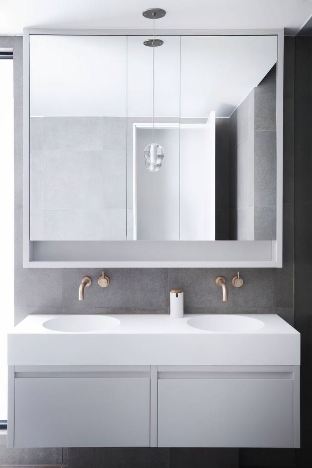 A contemporary double-vanity bathroom featuring a wall-mounted white vanity with integrated basins, brushed brass wall-mounted taps, grey tiled walls, and a large mirrored cabinet. Minimal pendant lighting and clean-lined cabinetry create a refined monochromatic scheme.
