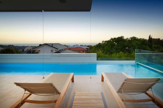 A modern rooftop or elevated pool terrace viewed through large frameless glass panels, with a bright blue infinity-style swimming pool, pale tiled paving, and two timber-framed lounge chairs facing the water. A timber-slatted side table sits in the foreground, while suburban rooftops, dense greenery, and a calm coastal-looking horizon are visible beyond the glass balustrade. The scene has a clean contemporary architectural character and a relaxed outdoor resort-like atmosphere.