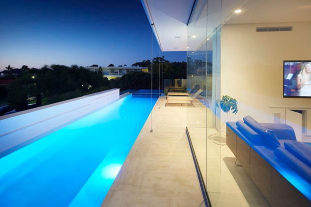 A contemporary rooftop or elevated pool terrace viewed beside a glass-walled interior. A long illuminated blue swimming pool runs along the left, bordered by pale stone paving and a white parapet. Full-height frameless glazing opens onto a minimalist lounge with a vivid blue sofa, television, recessed ceiling lighting, and neutral finishes. The image is taken at dusk, creating a bright, luxurious indoor-outdoor atmosphere.