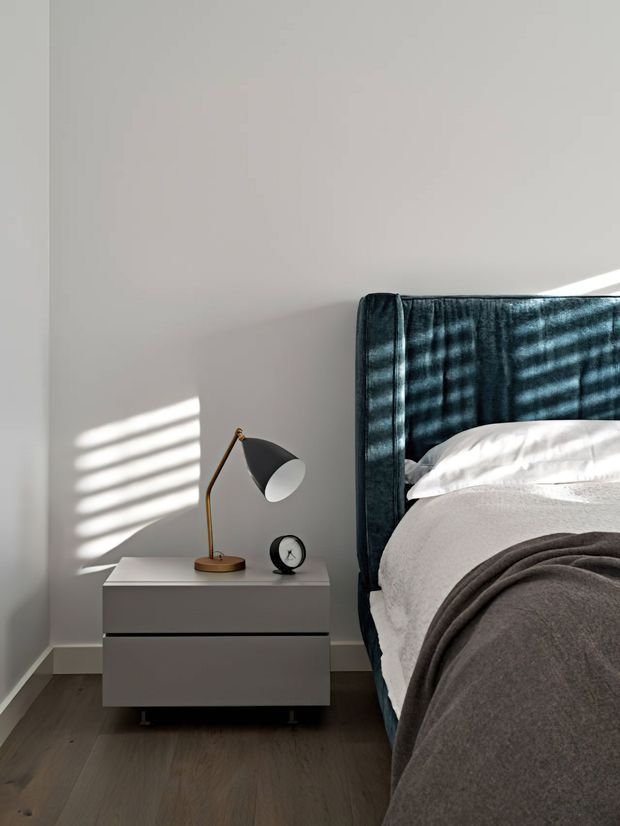 A softly lit contemporary bedroom vignette showing a dark teal upholstered bedhead, white bedding layered with a charcoal throw, and a minimalist white floating bedside table. A brass-and-black angled lamp and small black clock sit on the table, while sunlight filtered through blinds casts striped shadows across the pale wall and headboard. Dark timber flooring adds warmth and contrast.