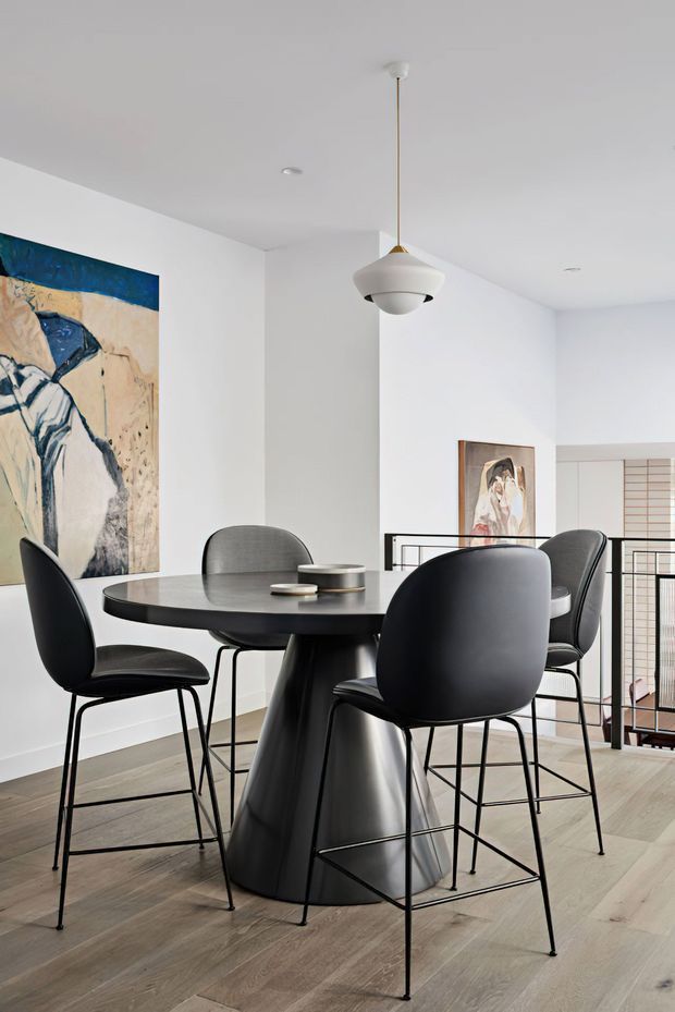 A bright, contemporary dining area with a dark round dining table and sculptural pedestal base, surrounded by black bar-height chairs. White walls, pale timber flooring, abstract artwork, a minimalist pendant light, and a black metal balustrade create a clean, refined interior.