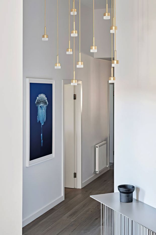 Contemporary interior hallway with tall light-grey walls, dark timber flooring, and a striking cluster of suspended pendant lights with brass-toned stems and white cylindrical shades. A framed blue artwork hangs on the wall, while a white console with vertically ribbed detailing and a small dark planter occupies the foreground. Open doorways and a radiator extend the corridor view.