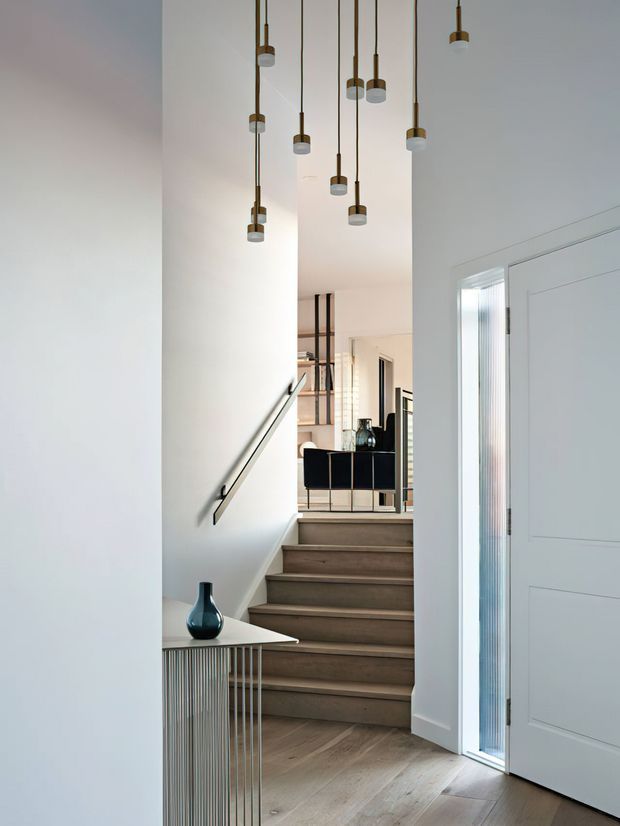 A bright, contemporary residential stairway with a tall void, pale timber flooring and stair treads, white walls, and a minimalist black handrail. Multiple slim pendant lights hang at varied heights above the stairwell. A fluted console with a dark decorative vase sits at the base, while a white door with vertical glazing opens on the right. A living area and dark furnishings are visible beyond the stairs.