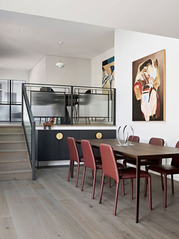 A refined open-plan dining area with a long dark timber dining table and burgundy upholstered chairs. Pale timber flooring, white walls and ceiling, black-framed glass and metal stair balustrades, and a dark fluted built-in cabinet create a crisp contemporary interior. Abstract artwork and sculptural decor add colour and visual interest, while the staircase and mezzanine-style glazing are visible in the background.