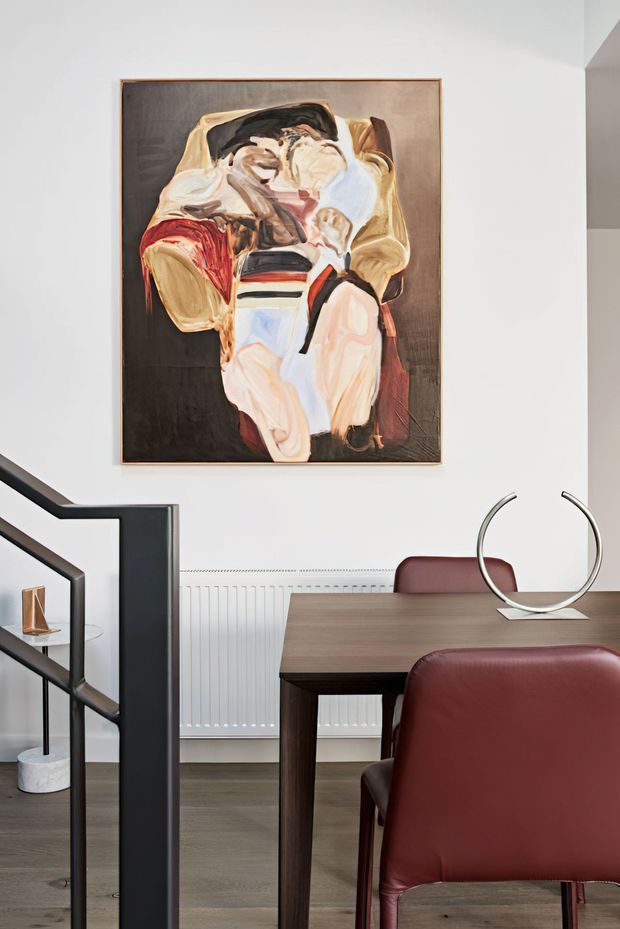 A refined dining area with a dark timber dining table and burgundy upholstered chairs, set against a white wall with a large expressive abstract painting. A sculptural silver circular ornament sits on the table, while a black metal stair balustrade and open shelving appear at the left. The room has timber flooring, a white radiator, and a restrained contemporary aesthetic with warm, muted tones.