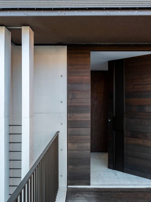 A close-up architectural view of a contemporary residential entryway. A dark timber-clad doorway with a large open pivot door sits beside smooth pale concrete walls and structural columns. A narrow metal balustrade and stair are visible in the foreground, while a perforated or mesh-like soffit and roof edge define the overhead structure. The palette is restrained, combining dark timber, pale concrete, and dark metal.