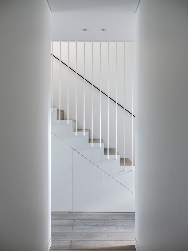 A minimalist white stairway viewed through a narrow hallway opening. The staircase has pale timber treads, white integrated cabinetry beneath, and a delicate vertical white balustrade with a slim black diagonal handrail. Pale grey timber flooring and recessed ceiling lights reinforce the clean, contemporary architectural composition.