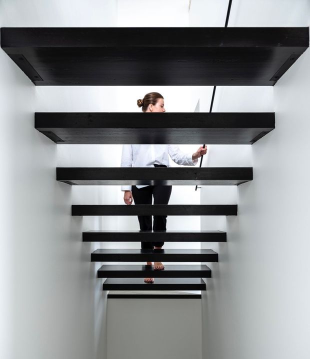 A minimalist interior view looking up a floating staircase composed of thick black horizontal treads, open risers, and concealed wall-mounted supports. White walls and bright natural light create a stark contrast, while a person is visible ascending the stairs. The design has a sculptural, contemporary character.