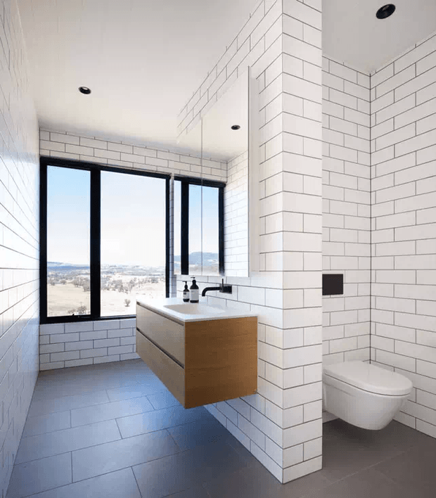Bright contemporary bathroom with white rectangular wall tiles, matching tiled ceiling, dark grey floor tiles, and a floating timber vanity with integrated basin. A wall-hung toilet sits behind a partial tiled partition. Black-framed full-height windows overlook a distant landscape, while recessed black ceiling lights and black tapware provide contrast.