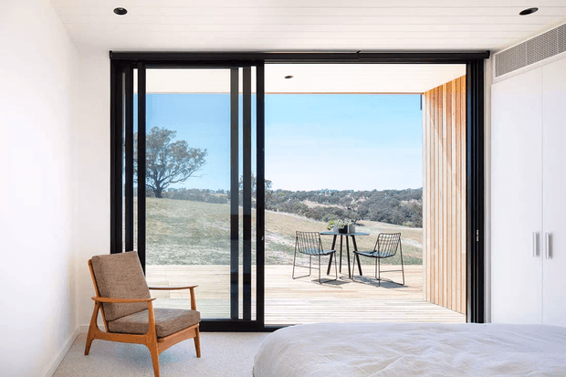 A bright contemporary bedroom with a low-profile bed, mid-century-style timber armchair, and expansive black-framed sliding glass doors opening onto a timber deck. The outdoor dining setting and rolling landscape are visible beyond, while vertical timber screening lines one side of the opening. The room has white walls, a white panelled ceiling, pale carpet, and integrated white cabinetry.