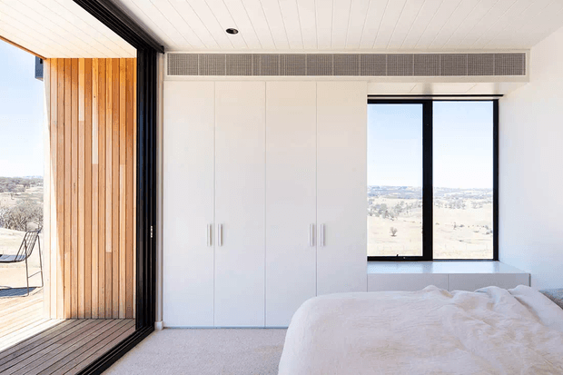 A bright, minimalist bedroom with a white built-in wardrobe, low-profile bed, pale carpet, and full-height black-framed glazing. Timber vertical cladding and decking are visible on the adjoining outdoor terrace, with an expansive landscape beyond. The room has a clean contemporary feel and abundant natural light.