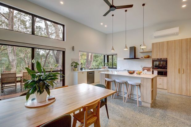 Bright open-plan kitchen and dining area with a large timber dining table, island bench with integrated seating, pale timber cabinetry, terrazzo-style flooring, pendant lights, and extensive windows overlooking trees. A ceiling fan and split-system air conditioner are visible, with a relaxed connection to the surrounding landscape.