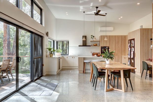 Bright open-plan kitchen and dining area with polished terrazzo flooring, extensive timber cabinetry, white tiled splashback, and a timber dining table. Large black-framed glazed doors and windows open to a leafy outdoor setting, while a ceiling fan, pendant lights, and indoor plants add functional and natural accents.