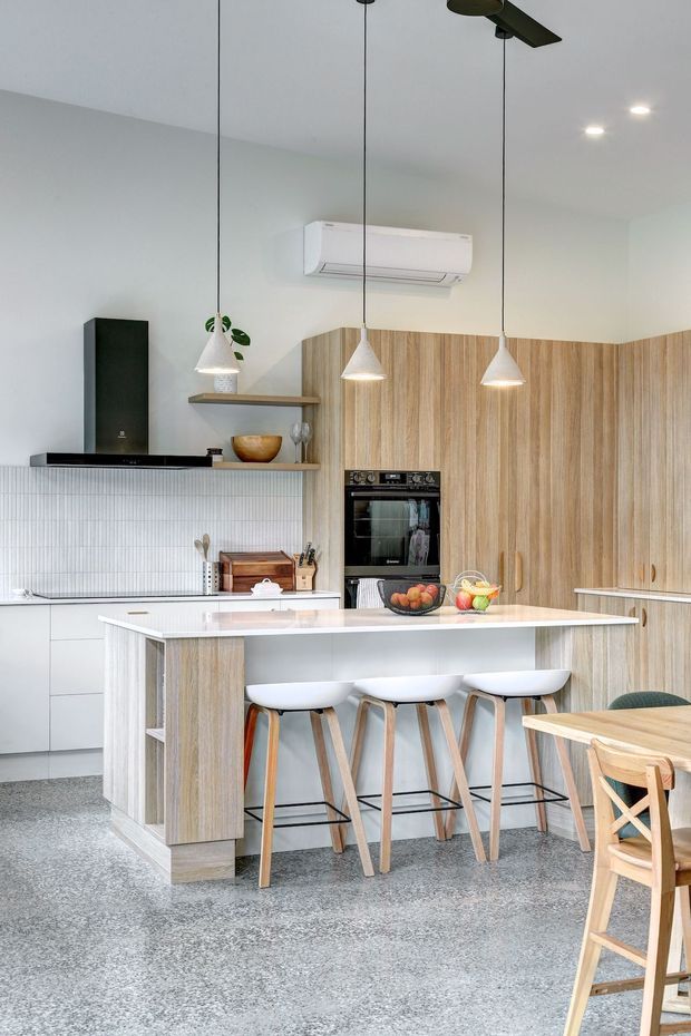 Bright contemporary kitchen featuring a large white island with integrated seating, pale timber cabinetry and wall panels, a white vertically tiled splashback, built-in black oven, open shelving, pendant lights, and a dining table with timber chairs. The room has a clean, airy feel with terrazzo-style flooring and neutral finishes.