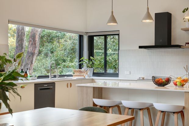 Bright contemporary kitchen with a pale island and integrated breakfast bar, white curved bar stools, light cabinetry, a white vertical-ribbed splashback, black-framed corner windows, pendant lights, and a dark rangehood. A timber dining table occupies the foreground, while leafy vegetation is visible outside the glazing.