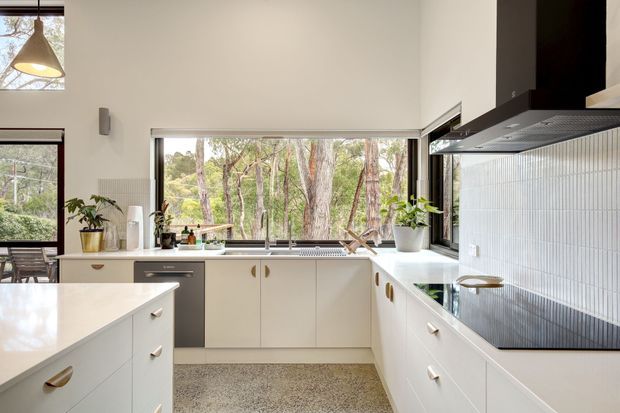 Bright contemporary U-shaped kitchen with white built-in cabinetry, a pale stone benchtop, white vertical tile splashback, dark integrated appliances, and a central island. Large windows provide views into a wooded landscape, while timber-toned handles, greenery, and a terrazzo-like floor add warmth. A sculptural pendant light hangs above the island.