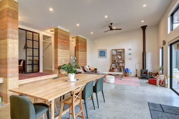 Open-plan living and dining space with a long timber dining table, upholstered dining chairs, exposed timber-clad structural columns, polished concrete flooring, a wood-burning stove, built-in shelving, ceiling fan, and expansive glazing. The room has a warm contemporary character with natural materials and abundant daylight.