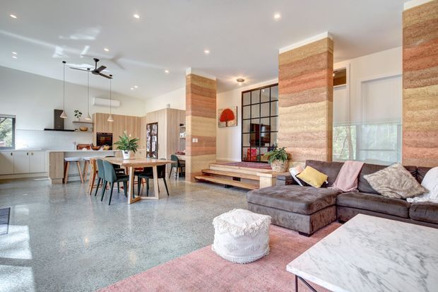 Bright open-plan living, dining and kitchen interior with polished concrete flooring, warm timber cabinetry, a large sectional sofa, and prominent textured, horizontally banded feature columns. Black-framed internal glazing, pendant lighting, ceiling fans and indoor plants contribute to a relaxed contemporary aesthetic.