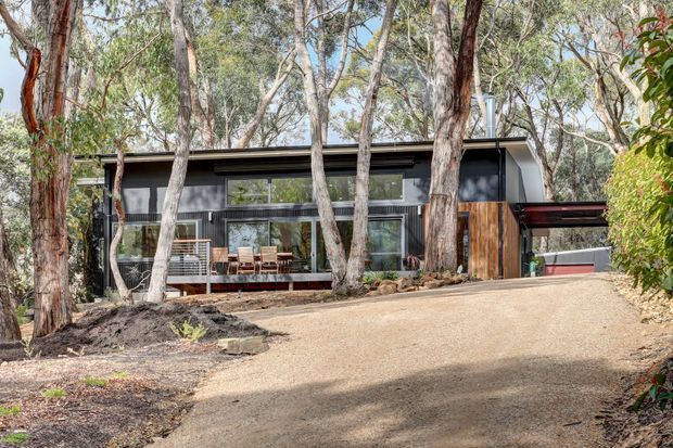 A contemporary two-storey dark-clad house nestled among mature eucalyptus trees, viewed from a broad landscaped driveway. The home features a low-pitched roof, extensive glazing, a timber-clad entry volume, and a covered carport or entrance structure.