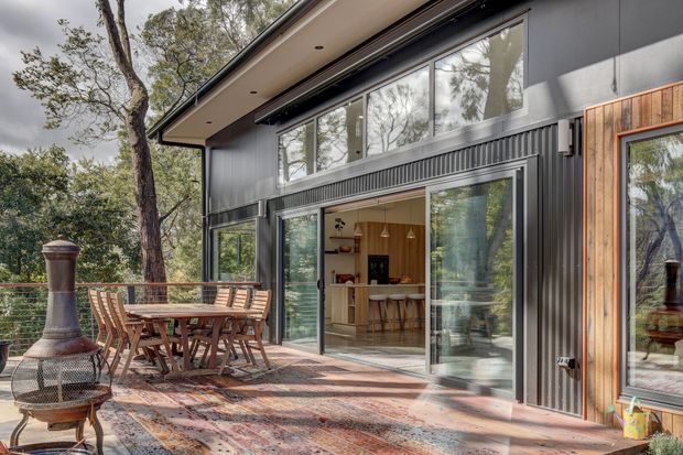 Exterior view of a contemporary, dark-clad house opening onto a furnished timber deck or patio. Large sliding glass doors reveal a warm kitchen and dining area inside, while vertical timber accents frame the facade. The outdoor area includes a dining table, chairs, patterned rug, and freestanding outdoor fireplace, set among mature trees.