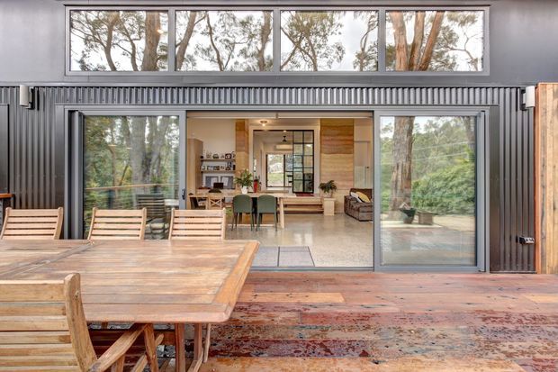 A contemporary indoor-outdoor living and dining space viewed from a timber deck. Large sliding glass doors open to a light-filled interior with a dining table, chairs, lounge seating, plants, and timber feature surfaces. Dark corrugated metal wall cladding surrounds the opening, while extensive glazing frames a leafy wooded setting.