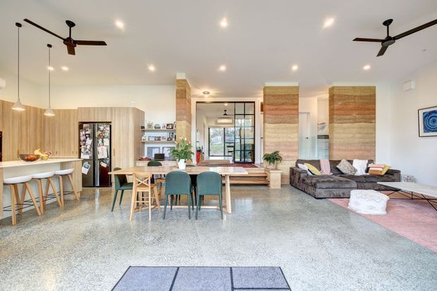 A bright, open-plan residential interior combining a kitchen, dining area and living room. The space features polished concrete flooring, light timber cabinetry, a dining table with upholstered chairs, a dark sectional sofa, exposed timber-and-plaster feature columns, large black-framed glazing, pendant lights and ceiling fans. Layered neutral and earthy textiles add warmth to the otherwise contemporary space.