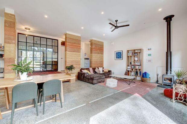 Bright open-plan living and dining area with polished concrete flooring, white walls, exposed layered stone or brick feature columns, black-framed industrial-style glazed partitions, a timber dining table, sectional seating, open shelving, a ceiling fan, and a freestanding wood-burning stove. The room has abundant natural light and a relaxed contemporary-industrial character.