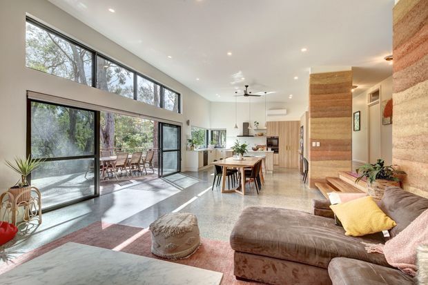 A bright, open-plan contemporary living, dining and kitchen area with extensive black-framed glazing, polished concrete flooring, timber cabinetry and a textured timber feature wall. The living room includes a large sectional sofa, while a dining table sits centrally and a compact kitchen extends along the rear. A covered outdoor dining area and leafy vegetation are visible through the glazing.
