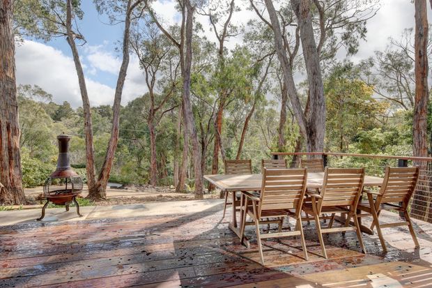 A timber outdoor deck dining area set among mature eucalyptus trees, with a large outdoor dining table and wooden chairs beside a metal balustrade. A freestanding chiminea-style outdoor fireplace stands at the edge of the deck, surrounded by bushland.
