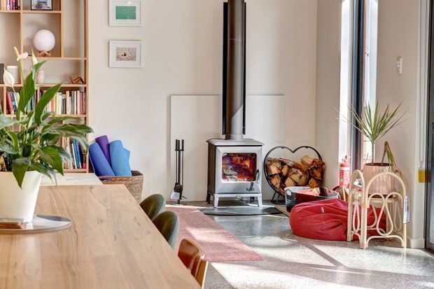 Bright, inviting open-plan living and dining area with a freestanding wood-burning stove, timber dining table, built-in shelving filled with books, indoor plants, colourful cushions and informal seating. Large glazing brings in abundant natural light, while a woven log basket and firewood reinforce the cosy character.