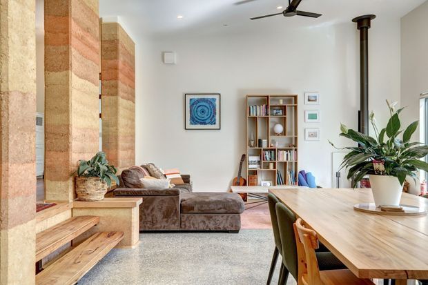 Bright open-plan living and dining area with a large sculptural stone fireplace and stepped timber hearth on the left, a brown sectional sofa, timber dining table with upholstered chairs, open bookshelf, indoor plants, and a black wood-burning flue. The room has white walls, polished concrete flooring, and a ceiling fan, with warm natural light and a relaxed contemporary feel.