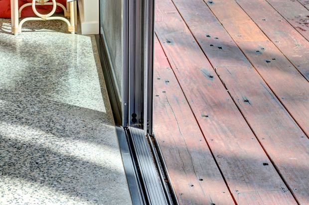 Close-up architectural detail of a threshold between an interior terrazzo floor and an exterior timber deck. A dark metal-framed glazed sliding door runs vertically through the centre, with sunlight casting shadows across the surfaces.