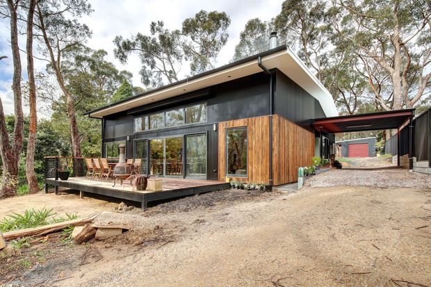 A contemporary single-storey home set among eucalyptus trees, featuring dark vertical cladding, warm timber wall panels, expansive glazing, a low-pitched roof with broad eaves, and a raised timber deck. A covered carport or sheltered passage connects toward a detached red-doored structure in the background.