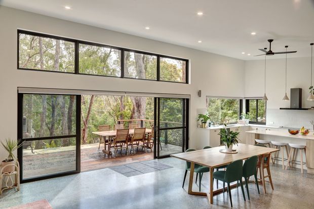 Bright open-plan dining and kitchen area with polished concrete flooring, extensive black-framed glazing, and direct access to a timber outdoor deck surrounded by trees. A timber dining table with green upholstered chairs occupies the foreground, while a contemporary white kitchen with island seating and pendant lights sits to the right. The room has a high ceiling, ceiling fans, abundant natural light, and a strong indoor-outdoor connection.