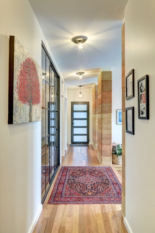A bright residential hallway and entry corridor with warm timber flooring, white walls, framed artwork, a patterned Persian-style runner rug, black-framed glazed internal doors, and a modern frosted-glass front door. A textured exposed brick or stone feature wall and indoor plants add visual interest, while circular ceiling lights illuminate the passage.