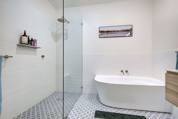 Bright contemporary bathroom with a frameless glass walk-in shower, freestanding white bathtub, white horizontal wall tiles, patterned geometric floor tiles, timber vanity cabinetry, and minimal wall-mounted fixtures.