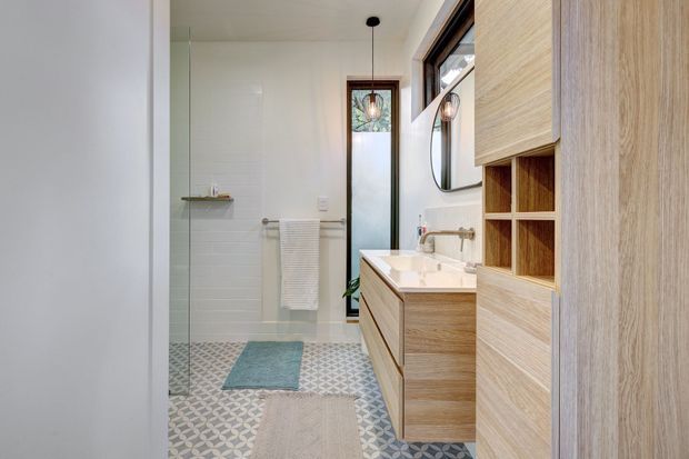 Bright contemporary bathroom featuring a walk-in glass shower, patterned geometric floor tiles, white tiled walls, a timber vanity with integrated basin, open shelving, and a tall narrow window. A round mirror and pendant light add decorative detail.