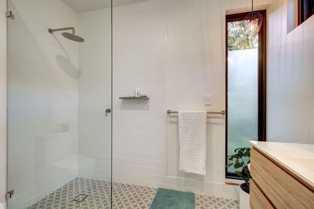 Bright contemporary bathroom with a frameless glass walk-in shower, round rainfall showerhead, white tiled walls, patterned geometric floor tiles, timber vanity cabinetry, and a narrow timber-framed window. A towel rail, basin, and small indoor plant complete the space.