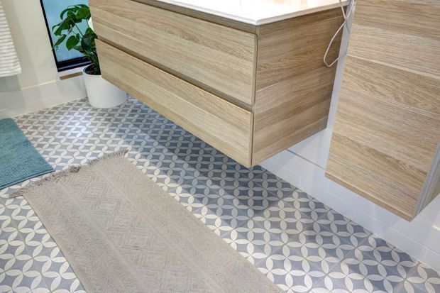 A contemporary bathroom vanity area with wall-mounted light wood cabinetry, a white benchtop, patterned geometric floor tiles in grey and white, and a pale textured runner. A partial white tiled wall and potted plant are visible.