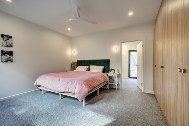 A bright, softly styled bedroom with a low upholstered bed, pink quilt, white and pale neutral bedding, bedside tables, wall-mounted reading lights, ceiling fan, carpeted floor, and extensive vertical timber wardrobe cabinetry along the right wall. An open doorway leads to a hall or adjoining space.