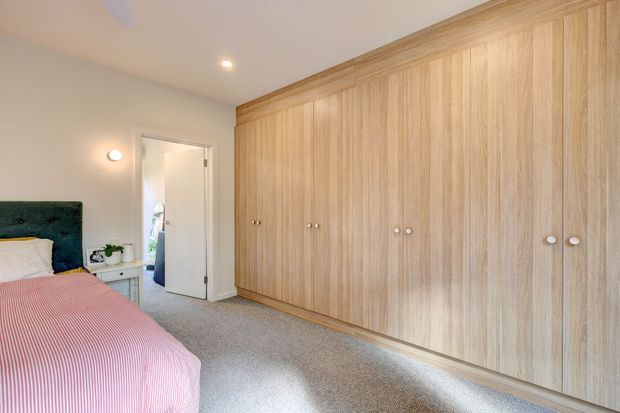 A bright bedroom with a large wall of built-in light timber wardrobes, grey carpet, a partially visible bed with a dark upholstered headboard and pink bedding, and a small bedside table. A doorway opens to an adjacent space.