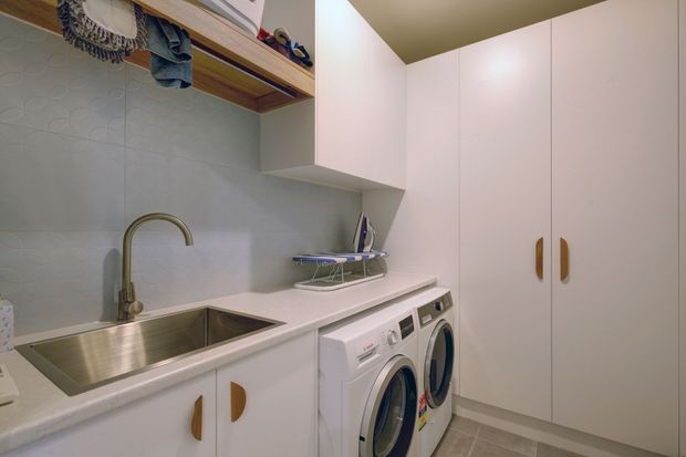 Compact, well-organised laundry room with white built-in cabinetry, a long pale benchtop, stainless-steel double sink, gooseneck tap, front-loading washer and dryer, tiled splashback, open timber shelving, and tall storage cupboards. The room has a clean, practical contemporary fit-out with warm timber handles and neutral finishes.