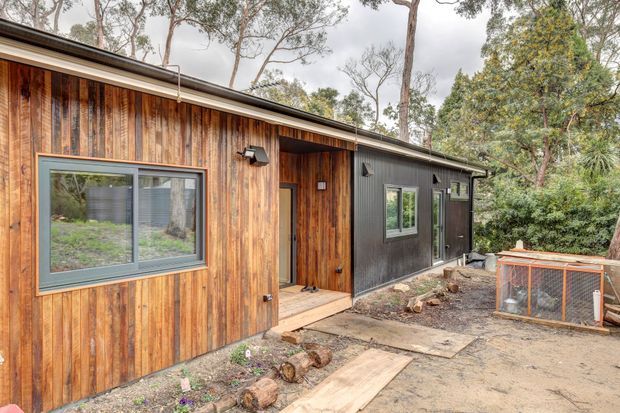 Exterior view of a compact single-storey dwelling in a wooded setting, combining vertical timber wall cladding with a dark corrugated-looking facade section, dark-framed windows, and a recessed entry porch. A simple low-pitched roof and informal timber-stepping pathway reinforce the natural bush setting.