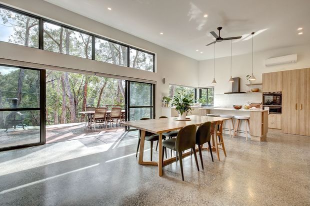 Open-plan dining and kitchen interior with polished concrete flooring, extensive black-framed floor-to-ceiling glazing, and a high white ceiling. A timber dining table with mixed chairs sits in the foreground, while a white kitchen with island seating, pendant lights, timber cabinetry, and integrated appliances occupies the right side. The glazing opens toward a leafy wooded outdoor setting and covered terrace.