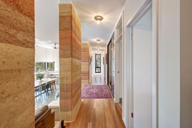 A bright residential hallway with warm timber flooring, white walls and trim, multiple doorways, and a patterned runner rug. A striking textured, horizontally banded earthen or stone feature wall separates the corridor from a visible dining area with glazing and greenery. Recessed ceiling lights and a glazed door or window at the far end add light and depth.
