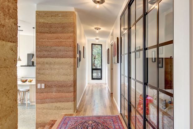 A bright residential hallway with pale timber flooring, a long black-framed glazed built-in storage wall on the right, and a textured horizontally banded plaster or earthen feature wall around a doorway on the left. A patterned runner adds colour, while the hallway leads to a glazed exterior door or window. A kitchen or dining area is partially visible through the opening on the left.