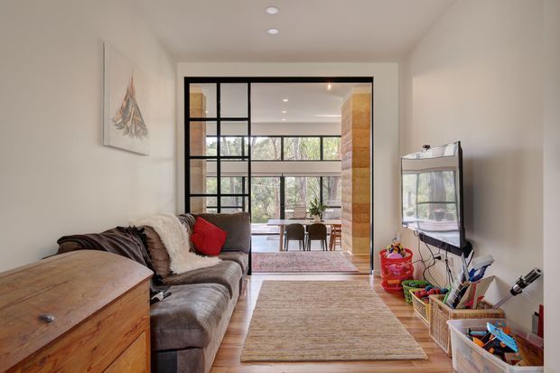 A bright, narrow living room with a large sectional sofa, timber chest, wall-mounted television, and a neutral rug. Black-framed glazed partition doors open toward a dining area with timber finishes and expansive garden-facing windows. The room has recessed ceiling lights, warm timber flooring, and a relaxed contemporary feel.