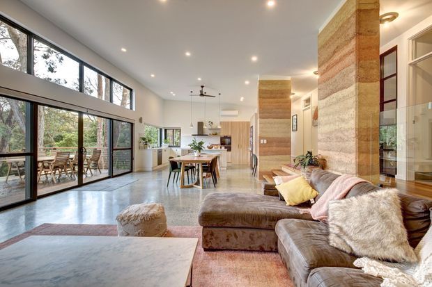 A spacious, open-plan living and dining area with polished concrete flooring, expansive black-framed glazing, a timber dining table, and a large sectional sofa. The room features tall natural-material feature columns, recessed ceiling lights, a ceiling fan, indoor plants, and views to a leafy outdoor dining area.