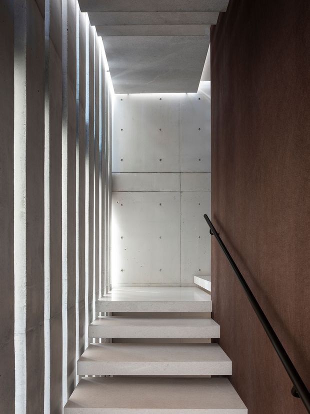 A narrow contemporary stairway with floating concrete steps, a smooth exposed concrete wall and ceiling, a dark brown feature wall, slim black handrail, and tall vertical glazing or slots along the left side admitting filtered light. The composition emphasizes strong linear geometry, material contrast, and a dramatic upward perspective.